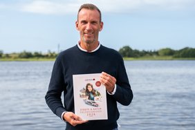 Ein Mann hält einen A-ROSA Katalog in die Kamera und steht dabei vor einem Fluss