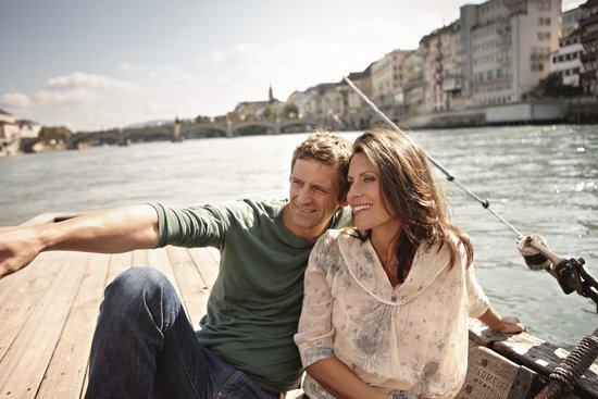Ein Paar sitzt entspannt auf einem Boot auf dem Rhein in Basel und genießt den Ausblick bei Sonnenschein. Im Hintergrund sind Gebäude am Ufer und eine Brücke.