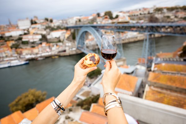 Zwei Hände halten ein Glas Portwein und ein Pastel de Nata vor der Ponte Dom Luís I Brücke in Porto, Portugal
