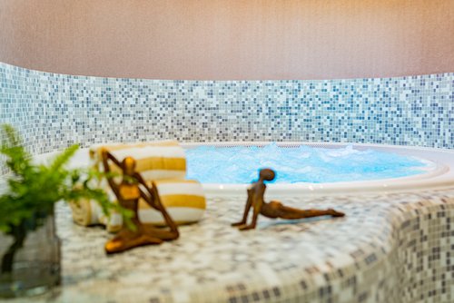 Innenwhirlpool mit blauen Wasserblasen, umgeben von Mosaikfliesen in Blau- und Grautönen, mit zwei gestreiften Handtüchern und zwei kleinen Bronze-Skulpturen in Yoga-Posen im Vordergrund