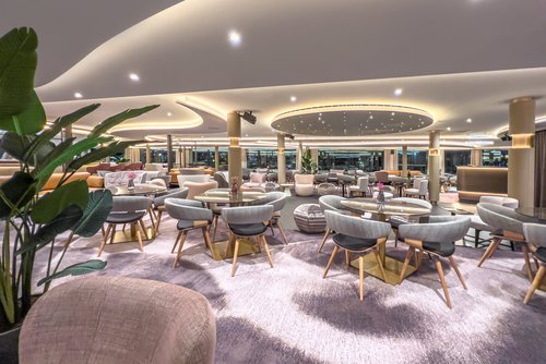 Moderne Lounge mit runden Tischen, grauen Stühlen mit Holzbeinen, Teppichboden und indirekter Deckenbeleuchtung