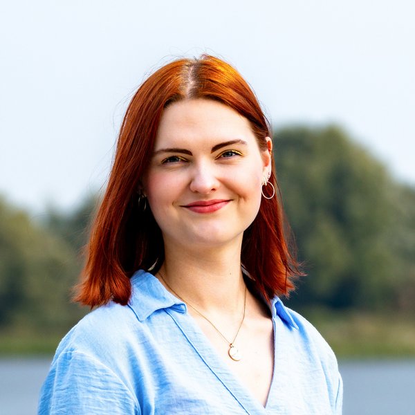 Nele Gaukel mit roten Haaren, blauem Hemd und Mondanhänger, lächelnd vor einem See und Bäumen im Hintergrund