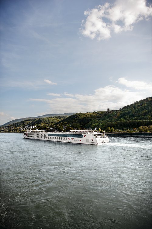 Ein Schiff der A-ROSA Rheinflotte gleitet am Nachmittag auf dem Rhein entlang, am Ufer grüne, bewaldete Hügeln und kleinen Siedlungen im Hintergrund.