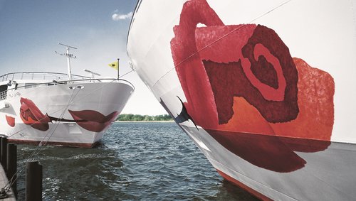 Nahaufnahme eines Flusskreuzfahrtschiffs der A-ROSA Flotte mit markanter Rose auf dem Bug im Vordergrund. Gegenüber steht ein zweites A-ROSA Schiff mit Blick auf den Bug.