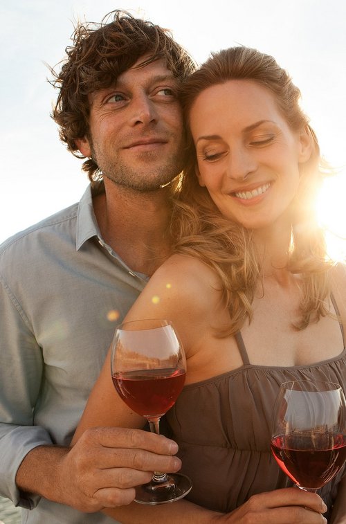 Ein Paar steht bei Sonnenuntergang mit einem Glas Wein in der Hand an der Rehling eines Flussschiffes. Im Hintergrund der Fluss und eine Uferlandschaft.