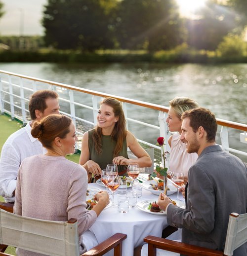 Yvonne Catterfeld und vier weitere Personen sitzen an einem gedeckten Tisch auf einem Sonnendeck am Wasser und genießen ein Essen bei Sonnenuntergang.