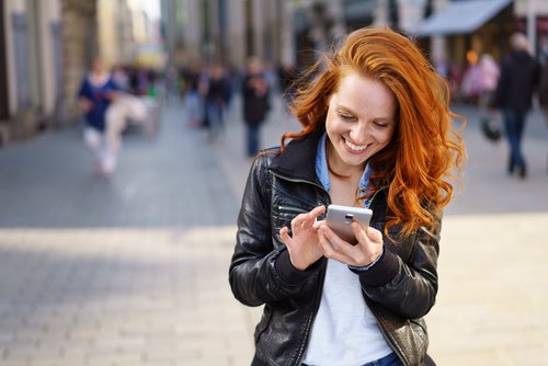 Eine rothaarige Frau tippt auf ihr Smartphone und lächelt während sie durch eine Stadt geht