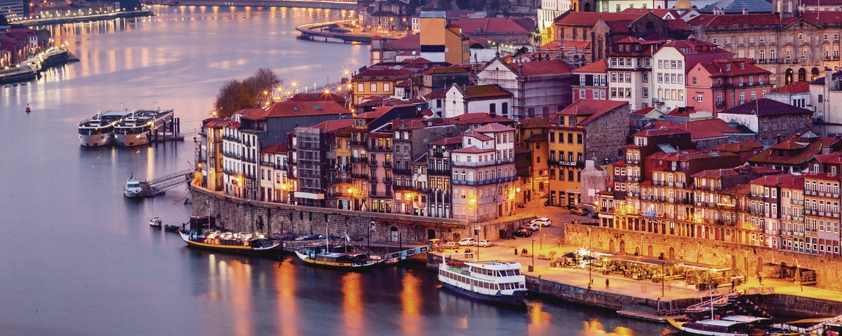 Abendliche Stadtansicht von Porto mit beleuchteten Gebäuden am Fluss Douro und der Arrábida-Brücke im Hintergrund.
