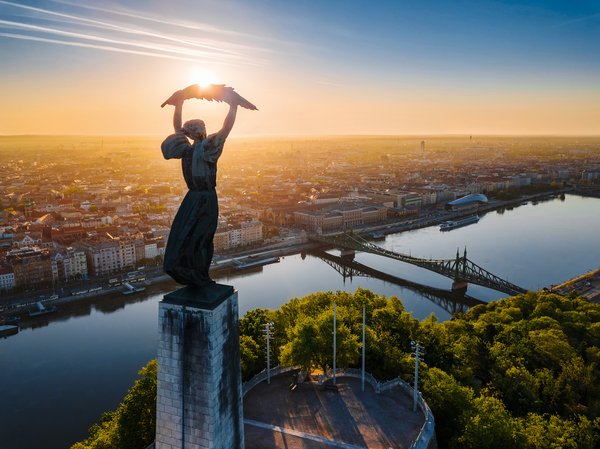 Die Freiheitsstatue in Budapest auf dem Gellértberg, umgeben von grünen Bäumen, mit Blick auf die Freiheitsbrücke über der Donau und der Stadt im Hintergrund bei Sonnenuntergang.