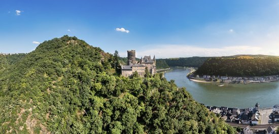 Panoramablick auf die mittelalterliche Burg Katz auf einem Hügel am Mittelrhein, umgeben von bewachsener Hügellandschaft und der Stadt St. Goarshausen am Flussufer.