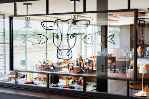 Das Restaurant SENA GRILL im industriell-modernen Design auf der A-ROSA SENA bei Tageslicht mit gedeckten Tischen und bodentiefen Fenstern mit Blick auf einen ruhigen Fluss.