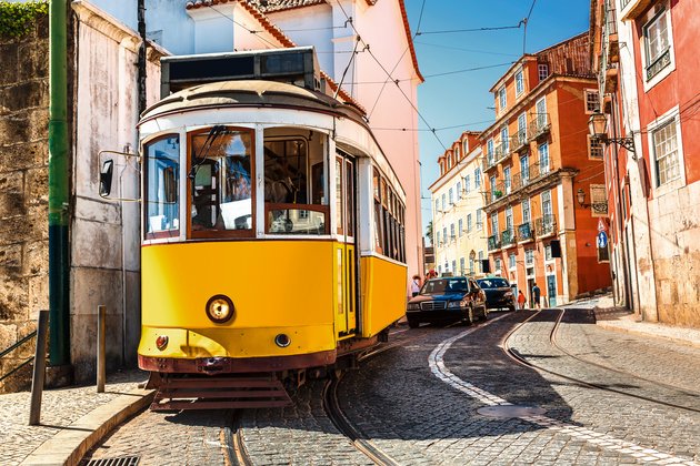 Gelbe Straßenbahn auf einer engen Kopfsteinpflasterstraße in Lissabon, umgeben von bunten Gebäuden und parkenden Autos.
