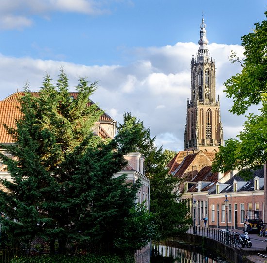 Ein Kanal in Utrecht, geziert von Bäumen und Gebäuden. Eine historische Kirche mit einem hohen Turm im Hintergrund spiegelt sich im Wasser.