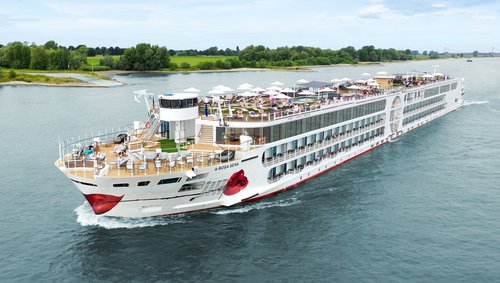 Die A-ROSA SENA fährt über den Rhein, das mit Liegestühlen und Sonnenschirmen ist gut besucht im Hintergrund das Ufer mit grüner Landschaft und Bäumen. 