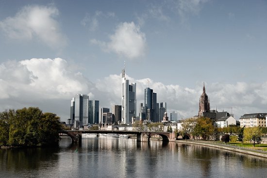 Frankfurter Skyline mit Hochhäusern unter bewölktem Himmel, mit dem Main und Brücke mit Parkanlage im Vordergrund.