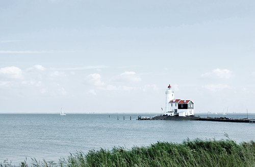 Ein Leuchtturmhaus am Ijsselmeer mit Schilf im Vordergrund und Segelbooten auf dem Meer, unter einem bewölkten Himmel.