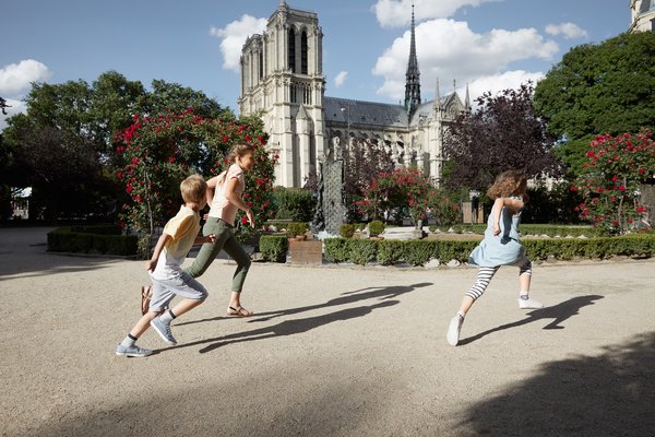 Drei Kinder rennen in einem Park mit blühenden Rosenbüschen vor der Kathedrale Notre-Dame in Paris bei sonnigem Wetter.