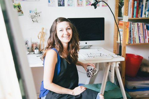 Die Illustratorin Tanja Meyer sitzt an Ihrem Schreibtisch, hält eine Tasse in der Hand und lächelt in die Kamera. Im Hintergrund stehen ein Computer und ein Bücherregal.