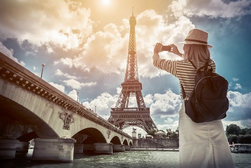 Touristin mit Strohhut und Rucksack fotografiert den Eiffelturm bei sonnigem Himmel von der Seine-Brücke in Paris.
