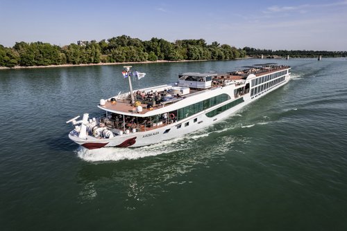 Die A-ROSA ALEA fährt auf dem Rhein an einem bewaldeten Ufer entlang. Das Sonnendeck ist mit Whirlpool, Sonnenliegen und Sonnensegeln ausgestattet.