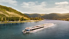 A-ROSA Flusskreuzfahrtschiff fährt auf einem breiten Fluss zwischen bewaldeten Hügeln unter bewölktem Himmel