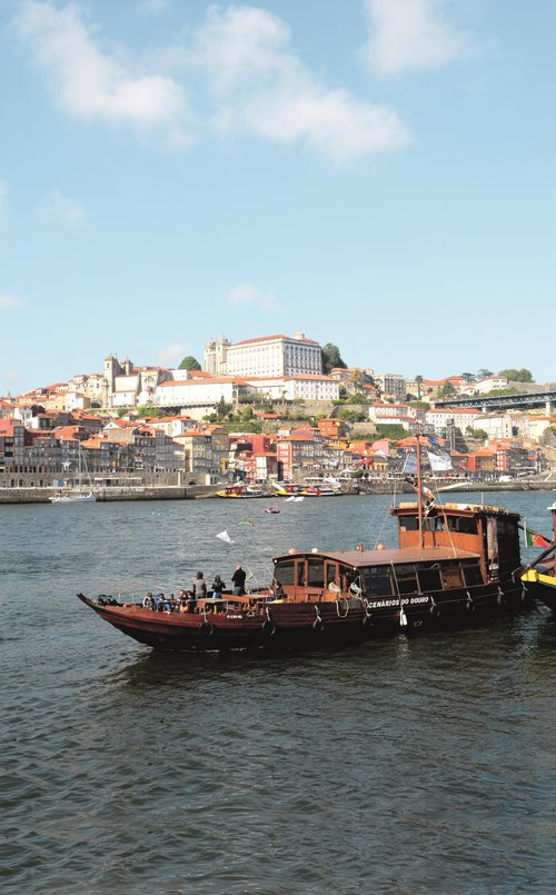 Traditionelles Holzboot auf dem Douro-Fluss vor der Stadt Porto mit bunten Häusern und historischen Gebäuden am Ufer.