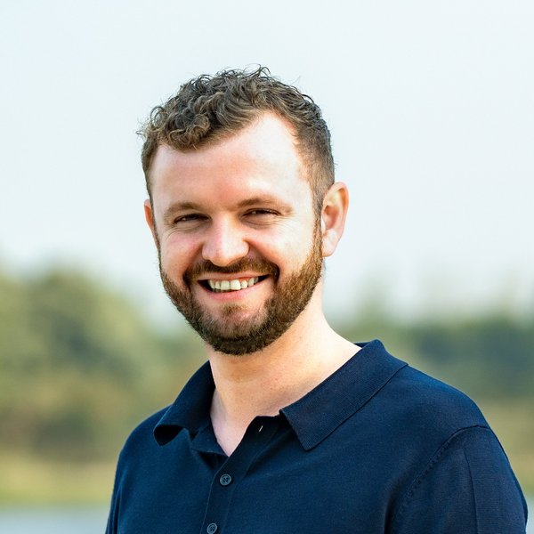 Hauke Heinrich lächelt, trägt ein dunkelblaues Poloshirt, steht vor unscharfem Naturhintergrund mit Wasser und Bäumen
