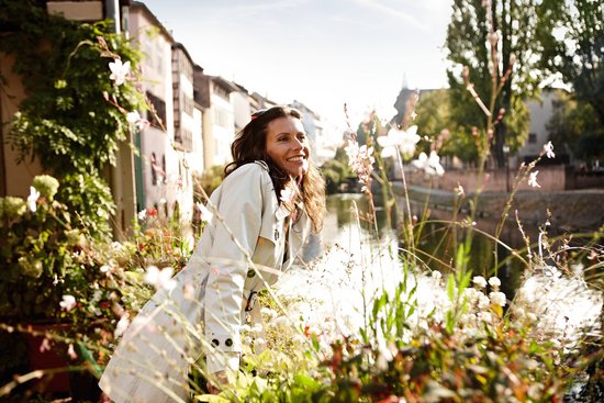 Eine Frau steht in Straßburg an einem Geländer zwischen Blumenkästen und schaut auf den Fluss, die Sonne scheint. 
