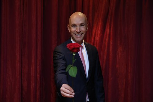 Michael Begasse steht lächelnd in einem eleganten Anzug vor einem Vorgang und hält eine rote Rose in der Hand.