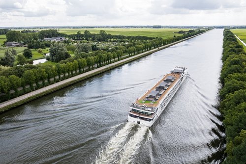 Ein Flusskreuzfahrtenschiff fährt auf dem Rhein am Ufer ein kleines Dorf, grüne Felder und Bäume. Das Sonnendeck ist mit Whirlpool, Sonnenliegen und Sonnensegeln ausgestattet.