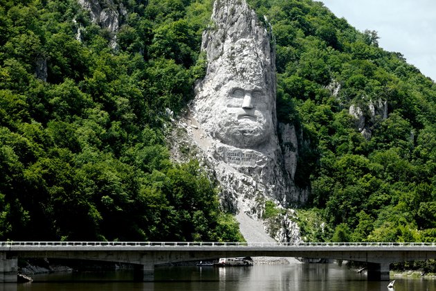 Felsenlandschaft am Donau-Delta mit Felsenstatue des Kopfes des Dakerkönigs Decebalus, umgeben von üppiger Vegetation mit einer Brücke und Fluss im Vordergrund.