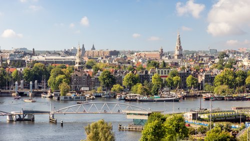 Blick auf Amsterdams Stadtzentrum mit Kanälen, historischen Gebäuden, grünen Bäumen und einer Fußgängerbrücke über das Wasser