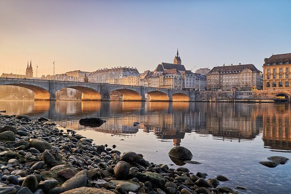 Steinbrücke über den Rhein mit historischen Gebäuden und Kirchturm in Basel bei Sonnenuntergang, Ufer mit Steinen im Vordergrund