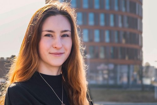 Junge Frau mit langen roten Haaren und schwarzem Pullover lehnt an einem Geländer vor einem modernen, runden Bürogebäude am Wasser.