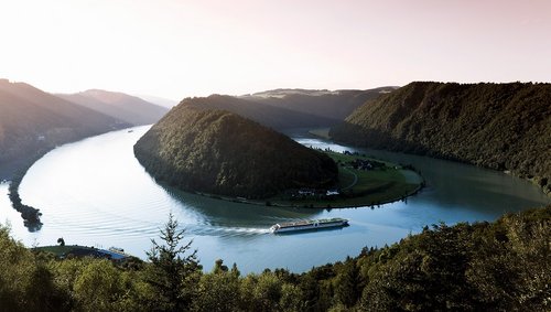 Ein Flusskreuzfahrtschiff der A-ROSA Flotte auf der Donau entlang der Schlögener Schlinge. Panoramaaufnahme mit Blick auf die angrenzende Hügellandschaft bei Sonnenaufgang.