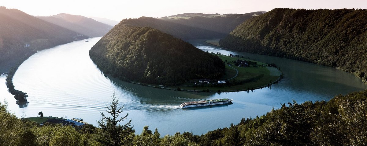 Ein Flusskreuzfahrtschiff der A-ROSA Flotte auf der Donau entlang der Schlögener Schlinge. Panoramaaufnahme mit Blick auf die angrenzende Hügellandschaft bei Sonnenaufgang.