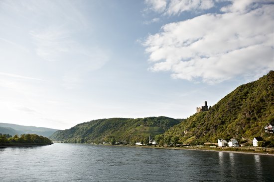 Blick auf die Loreley am Rhein mit dem Fluss, der sich durch die Landschaft schlängelt, umgeben von grünen Hügeln mit einer Festung im Hintergrund unter einem blauen, bewölkten Himmel.