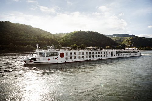 Ein Schiff der A-ROSA Rheinflotte gleitet am Nachmittag auf dem Rhein entlang, am Ufer grüne, bewaldete Hügeln und kleinen Siedlungen im Hintergrund.