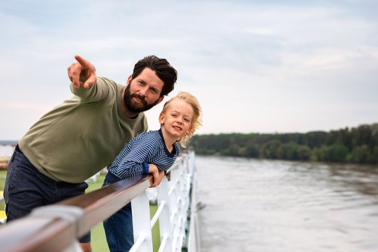 Vater und Sohn stehen auf dem Deck eines Flusskreuzfahrtschiffs und blicken über die Reling auf den Fluss. Der Vater zeigt auf etwas in der Ferne.