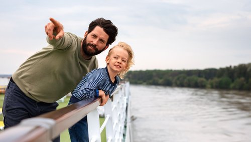 Vater und Sohn stehen auf dem Deck eines Flusskreuzfahrtschiffs und blicken über die Reling auf den Fluss. Der Vater zeigt auf etwas in der Ferne.