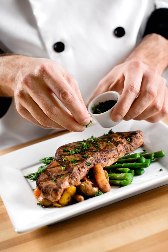 Koch streut frische Kräuter auf gegrilltes Steak mit grünen Bohnen und Gemüse auf weißem Teller.