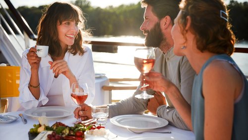 Drei Personen sitzen an einem gedeckten Tisch im Freien, trinken Wein und unterhalten sich bei Sonnenlicht.