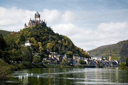 Blick auf die Reichsburg Cochem, umgeben von grünen Hügeln und Cochem an der Mosel mit Schwänen, unter einem bewölkten blauen Himmel.