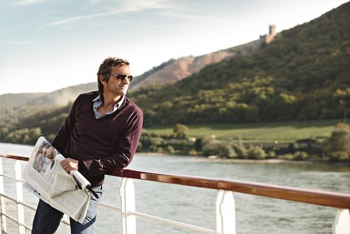 Ein Mann mit Sonnenbrille hält eine Zeitung auf dem Sonnendeck eines Flusskreuzfahrtschiffes bei Sonnenschein. Er genießt die Aussicht auf die Hügellandschaft am Rhein.