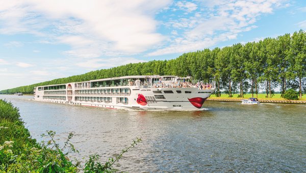 Die A-ROSA SENA fährt durch einen Rheinkanal an einer Baumreihe vorbei. Der Himmel ist blau und leicht bewölkt. 