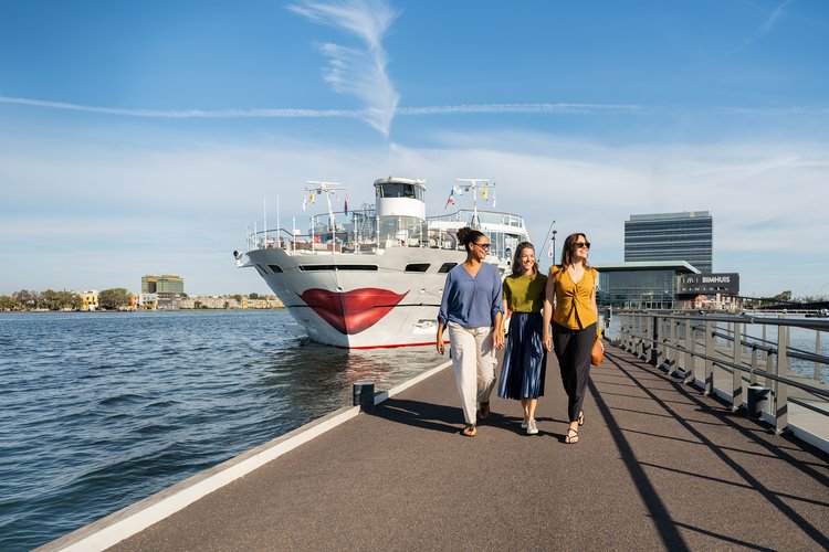 Drei Personen gehen auf einem Kai entlang, im Hintergrund ein Schiff mit einem großen roten Mund auf dem Bug und ein modernes Gebäude.