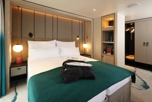 Eine stilvoll eingerichtete Suite auf der A-ROSA SENA mit Doppelbett, Sofa, Schreibtisch, begehbaren Kleiderschrank und bodentiefen Fenstern und Balkon bei Tageslicht. 