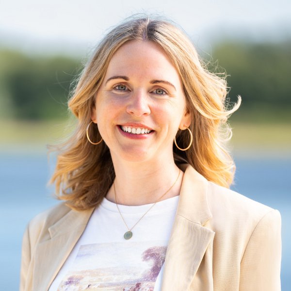 Annika Schmied mit blonden Haaren, beige Blazer, weißem T-Shirt und goldenen Creolen vor unscharfem Hintergrund mit Wasser