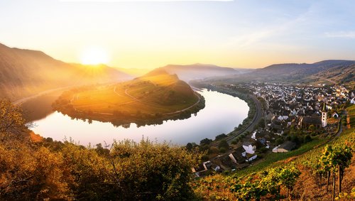 Eine Flussschleife der Mosel am Bremm Aussichtspunkt mit Blick auf Bremm bei Sonnenuntergang, umgeben von bewaldeten Hügeln und Weinbergen, unter einem klaren Himmel.