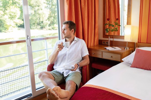 Mann in weißem Hemd und Shorts sitzt entspannt auf einem Stuhl am Fenster einer Kabine und trinkt Kaffee.